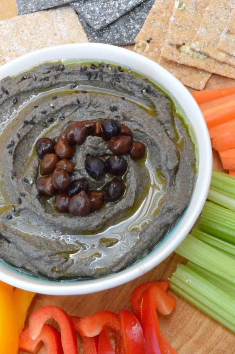 A bowl of black hummus topped with black chickpeas with crackers and crudités.