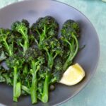 Bowl of lightly boiled purple sprouting broccoli with lemon wedge.