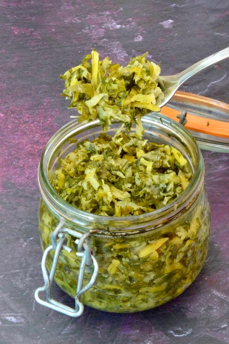 Forkful of homemade fermented kohlrabi with carrot tops taken from open jar.