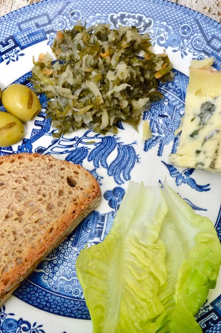 Kohlrabi and carrot top ferment on plate with cheese, olives, toast and lettuce.