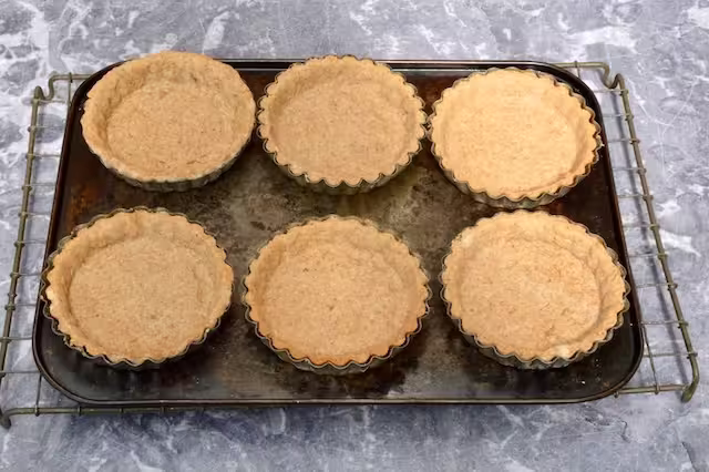 Six baked tart cases in tins on baking tray.