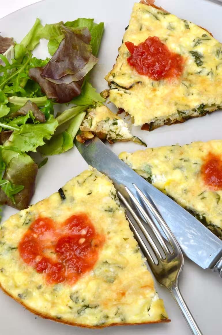 Courgette and ricotta frittata slices on plate with tomato sauce, salad and knife and fork.