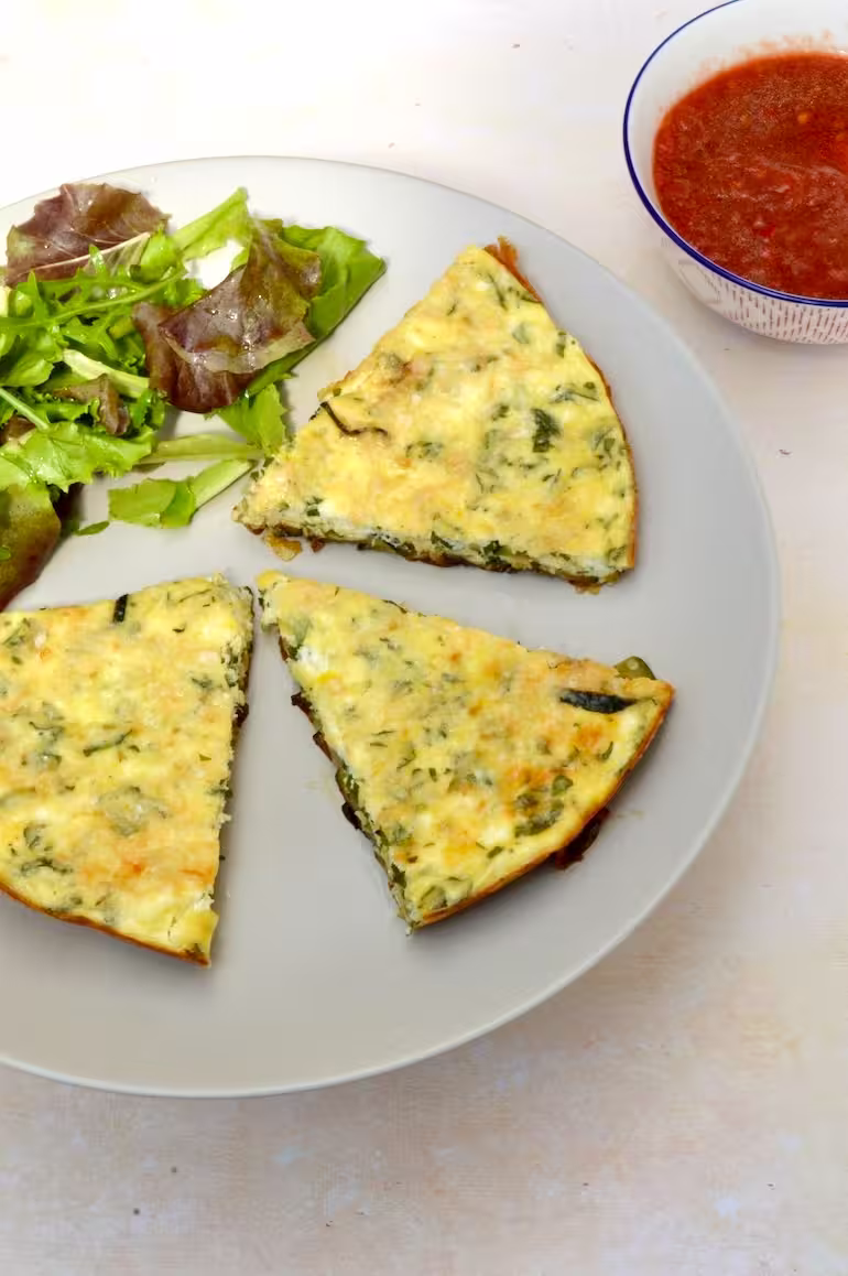 Three frittata slices on plate with salad and bowl of tomato sauce on side.