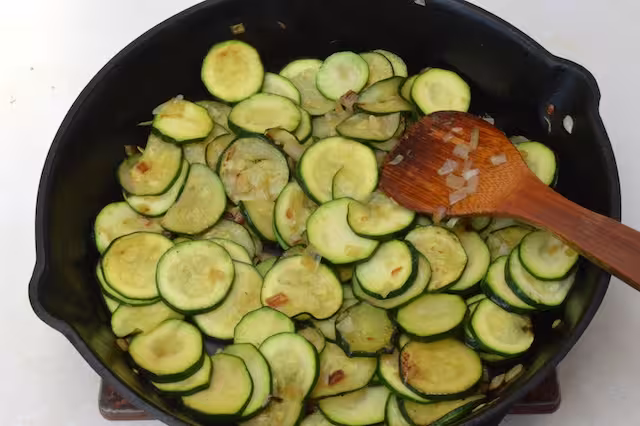 Courgettes frying in cast iron skillet with wooden spatula.