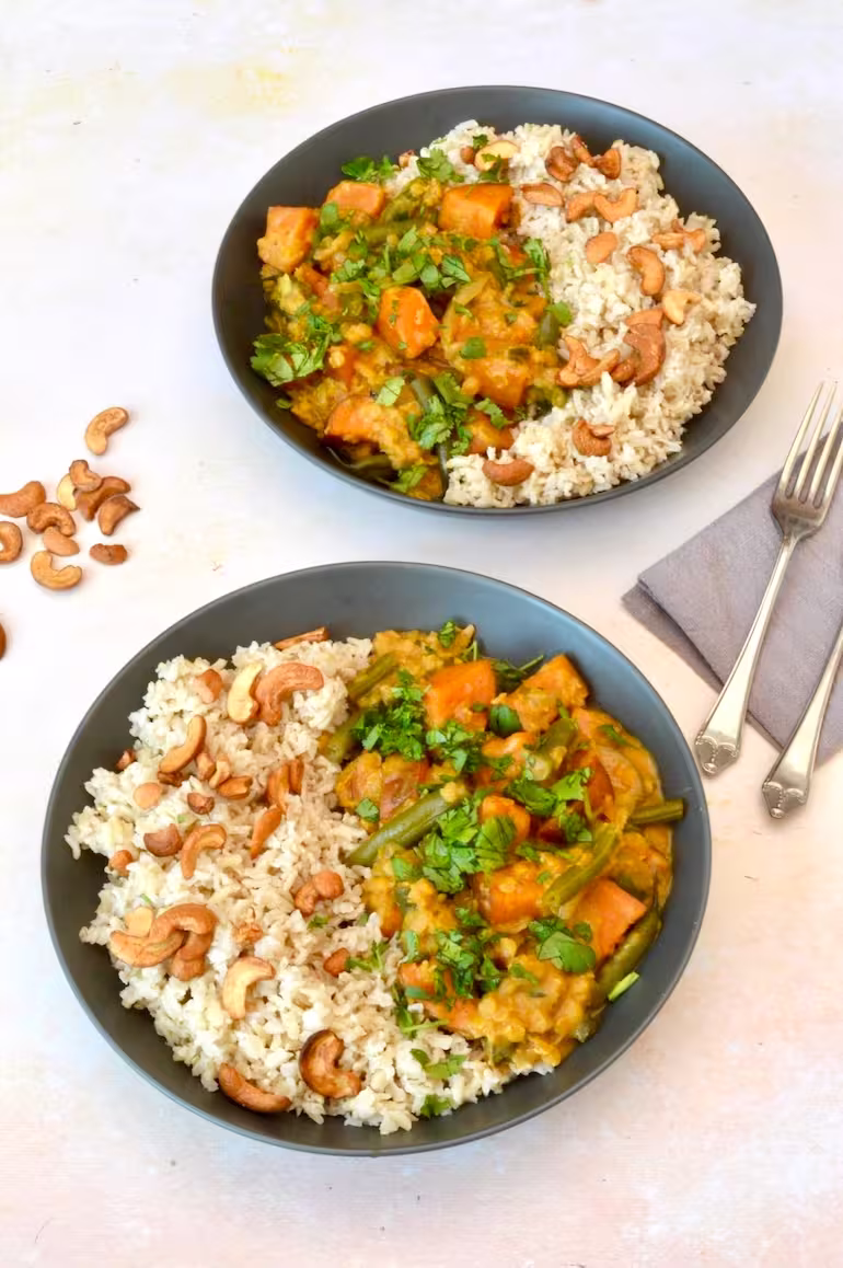 Two bowls of sweet potato and green bean curry with rice and cashew nuts.